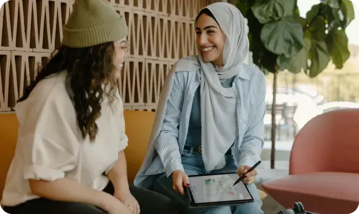 Two women sitting and smiling while discussing something on a tablet; one wears a hijab, the other a beanie